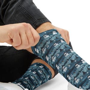 Puede incluir: Un par de calcetines con un patrón invernal repetitivo de muñecos de nieve blancos con sombreros y bufandas negras, intercalados con pinos de color verde oscuro sobre un fondo azul verdoso. Los calcetines se llevan sobre zapatos blancos.