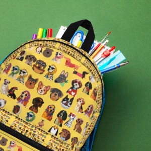 Puede incluir: Una mochila amarilla con un patrón repetitivo de varias razas de perros que llevan accesorios como gafas de sol, bufandas y jerséis. La mochila está abierta y muestra material escolar, incluidos bolígrafos de colores, una regla azul.