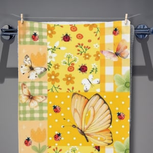 Puede incluir: Una toalla rectangular con un diseño de patchwork en tonos amarillos, verdes y blancos. El estampado incluye ilustraciones de mariposas naranjas y blancas, mariquitas rojas y motivos florales dispuestos en secciones de cuadros y lunares.