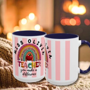 Puede incluir: Una taza de cerámica blanca con interior y asa azul oscuro, con un patrón de rayas verticales rosas y blancas. El diseño incluye un arcoíris colorido, una manzana roja y el texto: MISS OLIVIA TEACHER you make a difference.