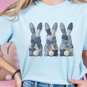 Puede incluir: Camiseta azul claro con tres diseños de conejos de patchwork de mezclilla. Cada conejo tiene un lazo y una cola blanca en forma de flor. Los conejos están hechos de varios patrones de mezclilla, incluyendo floral y cuadros.