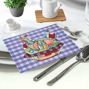Puede incluir: Un mantel individual con forma de gallina de retazos sobre un fondo a cuadros morado y blanco. El mantel individual presenta varios patrones y colores de tela, incluyendo rojo, verde, azul y amarillo. Una taza de té, un platillo y una tetera blancos están en el fondo.
