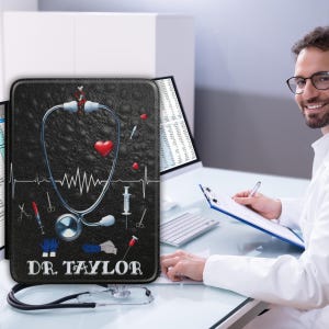 Puede incluir: Una funda negra para tableta con temática médica con un diseño de estetoscopio, corazón y herramientas médicas. La funda tiene el texto "DR. TAYLOR". Un estetoscopio está en primer plano.