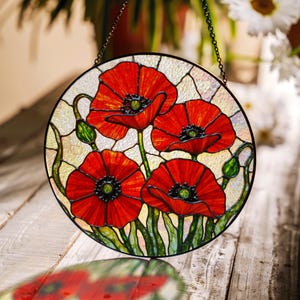 Op de afbeelding: Ronde glas-in-lood raamkunst met levendige rode klaprozen met groene stelen en bladeren. De klaprozen hebben zwarte centra en zijn geplaatst tegen een crèmekleurige achtergrond. Een decoratieve ketting is bevestigd om op te hangen.