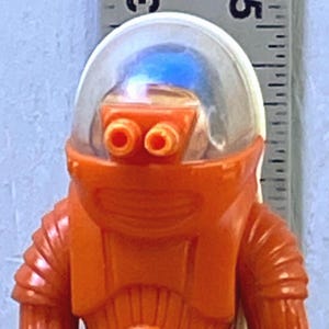 May include: Orange vintage toy robot figure with a clear helmet and silver accents. The robot has a detailed design and stands upright. Two small circular tokens are at the base of the robot.