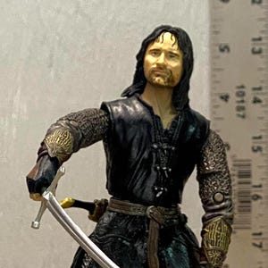 May include: A male action figure with long dark hair, wearing a black tunic over chainmail, dark trousers, and boots. The figure holds a silver sword in the right hand. A ruler in the background shows the figure is approximately 6.5 inches tall.