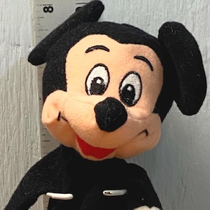 Puede incluir: Mu&ntilde;eco de peluche de Mickey Mouse, con cuerpo negro, pantalones cortos rojos con botones blancos, guantes blancos y zapatos amarillos. El mu&ntilde;eco tiene una cara sonriente con orejas negras y boca roja. El mu&ntilde;eco mide unos 20 cm de alto.