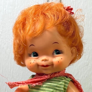 Könnte beinhalten: Vintage Puppe mit rotem Haar, blauen Augen und Sommersprossen. Die Puppe trägt ein grün-weiß gestreiftes Kleid, einen roten Schal und eine cremefarbene Hose. Zwei kleine Spielsteine aus Holz liegen zu ihren Füßen.