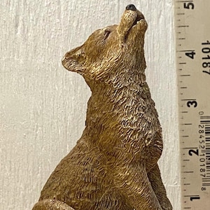 May include: A detailed bronze-colored wolf figurine in a seated position, head tilted back as if howling. The sculpture sits on a textured, dark brown base. Two small, round game pieces are in the foreground. A ruler is in the background.