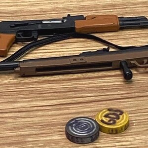 May include: Two miniature toy rifles with black and brown plastic components, including a black sling attached to one rifle. Two small circular tokens are positioned below the rifles, one yellow with black text and one dark gray with a circular pattern.