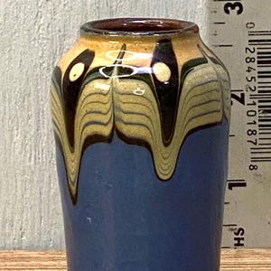 May include: A small, handcrafted ceramic vase with a blue body and a decorative top. The top has a brown rim and a yellow and brown pattern. Two small, round ceramic pieces are at the base of the vase.