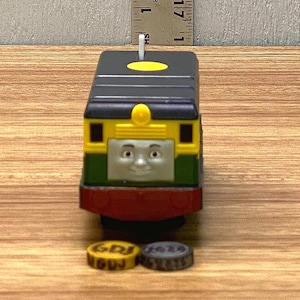 Könnte beinhalten: Eine Spielzeug-Lokomotive mit einem lächelnden Gesicht, bemalt in Gelb, Grün, Schwarz und Rot. Der Motor hat eine kleine weiße Antenne und steht auf einer Holzoberfläche. Zwei kleine, runde Metallobjekte befinden sich vor dem Zug.
