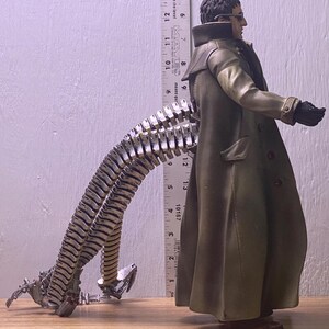 May include: A detailed action figure of Doctor Octopus, featuring a long olive green coat, black gloves, and metallic tentacles. The figure is posed with one arm extended. A ruler is placed next to the figure, measuring approximately 13 inches.