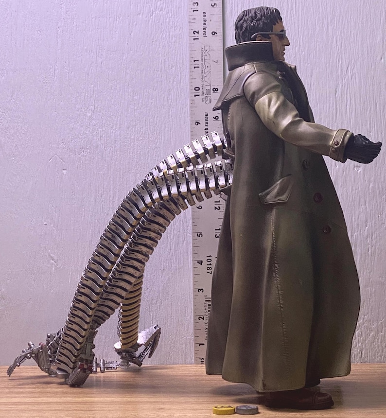 May include: A detailed action figure of Doctor Octopus, featuring a long olive green coat, black gloves, and metallic tentacles. The figure is posed with one arm extended. A ruler is placed next to the figure, measuring approximately 13 inches.