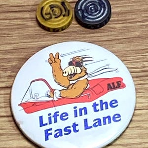 May include: A round white pinback button featuring a cartoon character wearing sunglasses and driving a red speedboat, with the text "ALF" and "Life in the Fast Lane" in blue, accompanied by two small circular game tokens in gold and black.