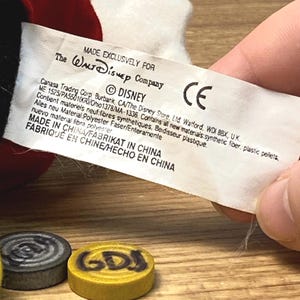 Puede incluir: Una etiqueta blanca con texto negro, sostenida por una mano, dice "MADE EXCLUSIVELY FOR The Walt Disney Company &copy; DISNEY CE MADE IN CHINA". Dos peque&ntilde;as piezas de juego redondas est&aacute;n en primer plano, una gris y otra amarilla.