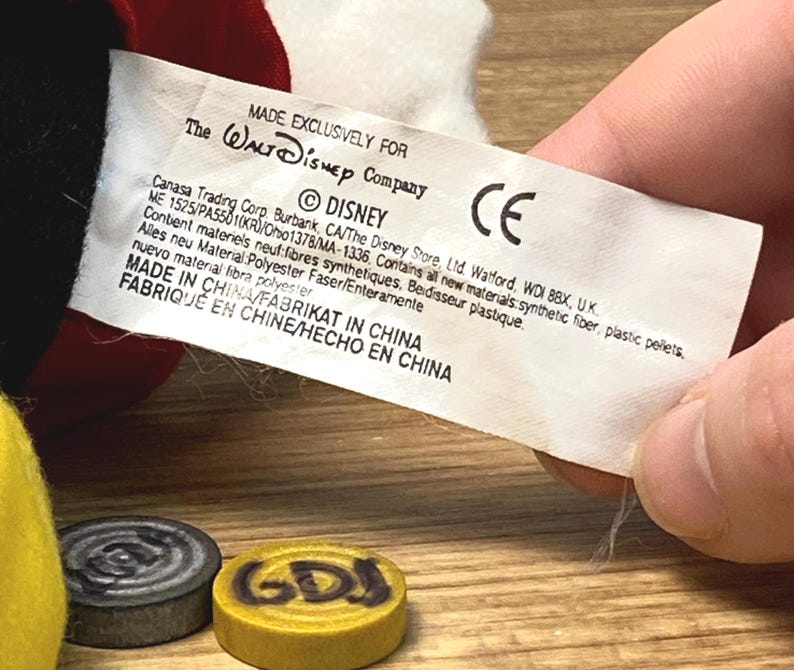 Puede incluir: Una etiqueta blanca con texto negro, sostenida por una mano, dice "MADE EXCLUSIVELY FOR The Walt Disney Company &copy; DISNEY CE MADE IN CHINA". Dos peque&ntilde;as piezas de juego redondas est&aacute;n en primer plano, una gris y otra amarilla.