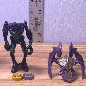 Peut inclure: Quatre figurines d'extraterrestres aux couleurs noir, vert et violet sont présentées sur une surface en bois. Les figurines ont des designs variés, avec des caractéristiques d'insectes et d'humanoïdes. Une règle est en arrière-plan, et deux petits jetons sont au premier plan.
