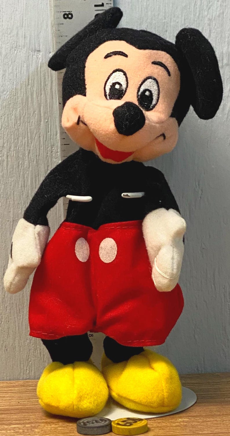 Puede incluir: Mu&ntilde;eco de peluche de Mickey Mouse, con cuerpo negro, pantalones cortos rojos con botones blancos, guantes blancos y zapatos amarillos. El mu&ntilde;eco tiene una cara sonriente con orejas negras y boca roja. El mu&ntilde;eco mide unos 20 cm de alto.