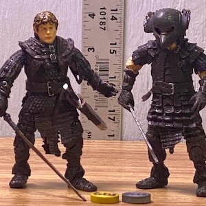 May include: Two detailed action figures of characters in dark armor, one with a staff and the other with a sword. The figures are standing next to a ruler, with two small, round, yellow and gray objects at their feet. The figures are approximately 3 inches tall.