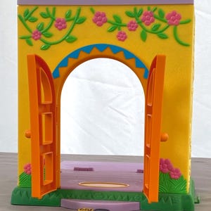 May include: A colorful toy playset featuring a yellow archway with orange doors and a purple roof. The archway is decorated with pink flowers and green vines. The base is green and purple, with a small coin on the floor.