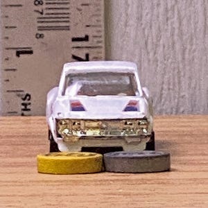Puede incluir: Un coche de juguete blanco con detalles azules y rojos en el capó y una parrilla cromada. Dos neumáticos de juguete, uno amarillo y otro gris, están delante del coche. Una regla está en el fondo.