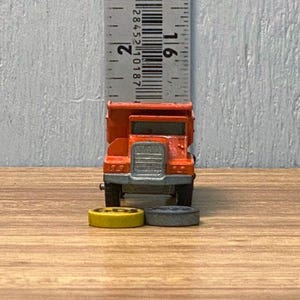 Pode incluir: Um caminhão basculante de brinquedo laranja com uma grade prateada e rodas pretas. O caminhão está posicionado em frente a uma régua, com duas pequenas rodas de brinquedo na frente. O caminhão tem cerca de 5 cm de comprimento.