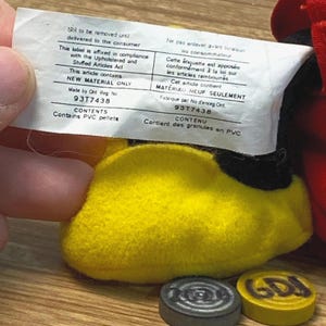 Puede incluir: Primer plano del pie amarillo de un juguete de peluche con suela negra. Una etiqueta blanca con texto en ingl&eacute;s y franc&eacute;s se sostiene encima. Dos peque&ntilde;as fichas redondas de madera en la parte inferior.
