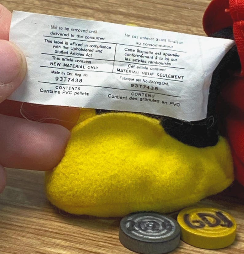 Puede incluir: Primer plano del pie amarillo de un juguete de peluche con suela negra. Una etiqueta blanca con texto en ingl&eacute;s y franc&eacute;s se sostiene encima. Dos peque&ntilde;as fichas redondas de madera en la parte inferior.