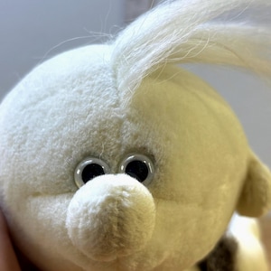 May include: Close-up of a soft toy with a round, off-white body and a rounded nose. The toy has large, shiny black eyes and a tuft of white hair. The toy's design is simple and child-friendly.