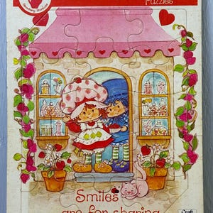 May include: A vintage Strawberry Shortcake puzzle featuring a pink awning, heart accents, and the text "Smiles are for sharing." The puzzle depicts Strawberry Shortcake and a friend, with a cat and potted plants. For ages 4 to 7.