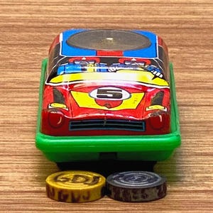 Könnte beinhalten: Ein Oldtimer-Rennwagen-Spielzeug in Rot, Gelb, Blau und Schwarz. Das Auto hat die Nummer "5" und steht auf einer grünen Basis. Zwei runde Spielmarken, eine gelbe und eine graue, befinden sich vor dem Auto.