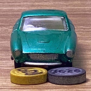 Puede incluir: Un coche de juguete de color verde azulado con una parrilla y faros plateados. Dos pequeñas fichas de juego redondas están delante del coche: una amarilla con un diseño marrón y otra gris con un diseño de círculos concéntricos. Sobre una superficie de madera.