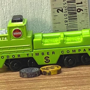 Könnte beinhalten: Ein grüner Spielzeug-Waggon mit dem Text "SODOR TIMBER COMPANY" und der Nummer "8374". Der Waggon hat schwarze Räder und eine rote und silberne Kupplung. Zwei kleine Holzscheiben liegen vor dem Waggon.