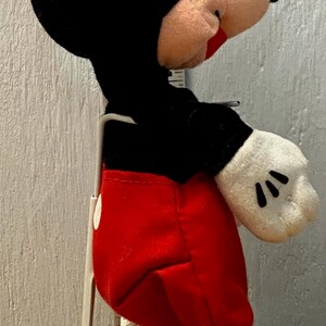 Puede incluir: Un peluche de Mickey Mouse, con cabeza y orejas negras, cara rosa y pantalones cortos rojos. El juguete tiene guantes blancos con detalles negros y zapatos amarillos. Est&aacute; montado sobre un soporte met&aacute;lico.