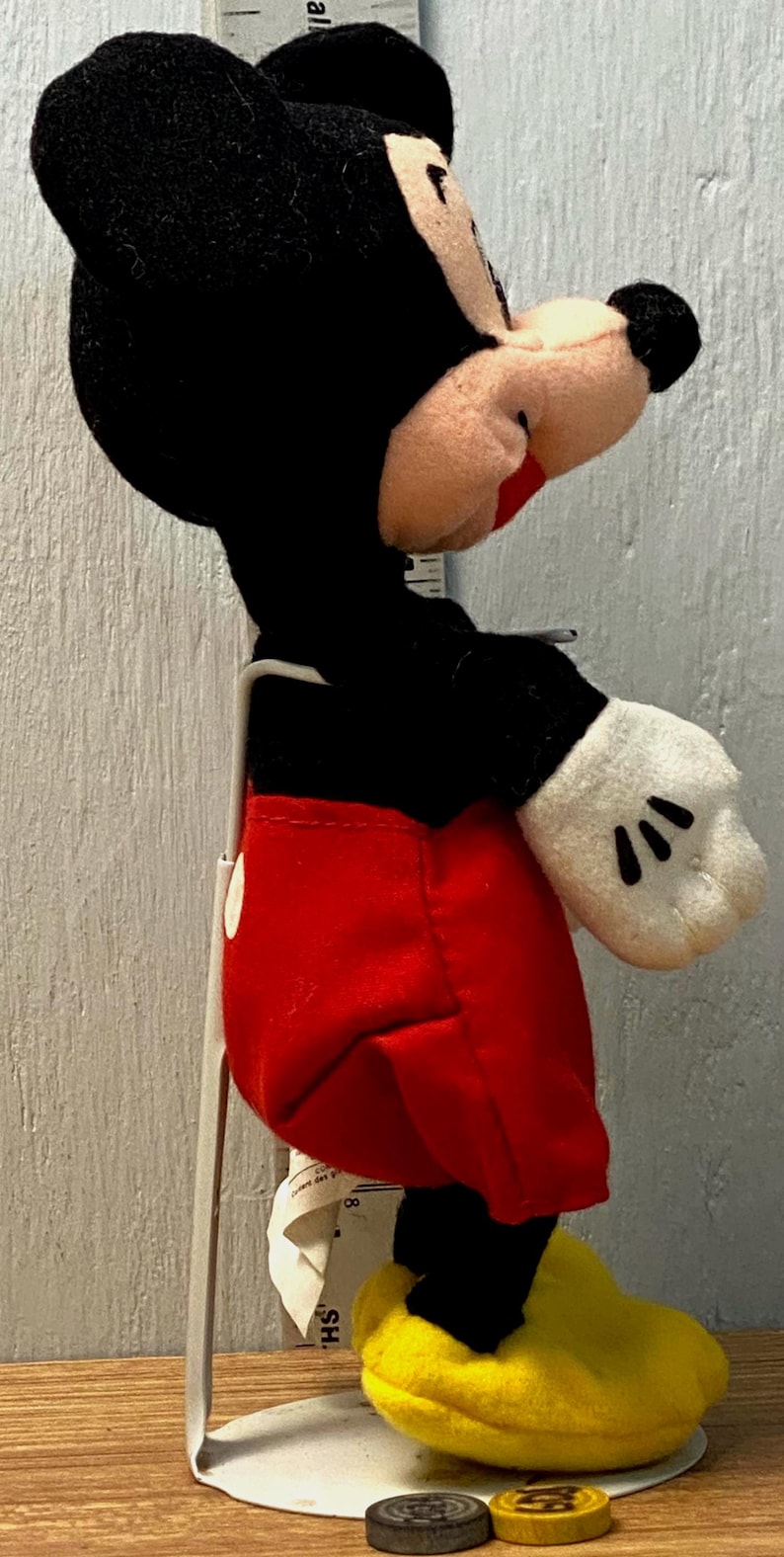 Puede incluir: Un peluche de Mickey Mouse, con cabeza y orejas negras, cara rosa y pantalones cortos rojos. El juguete tiene guantes blancos con detalles negros y zapatos amarillos. Est&aacute; montado sobre un soporte met&aacute;lico.