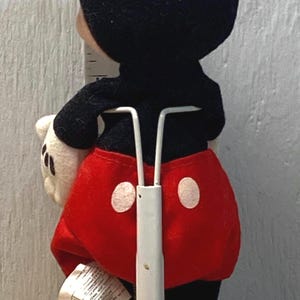 Puede incluir: Un peluche de Mickey Mouse, visto desde atr&aacute;s, con orejas negras, cuerpo negro, pantalones cortos rojos con botones blancos y zapatos amarillos. El juguete est&aacute; sobre un soporte blanco. Una regla es visible en el fondo.