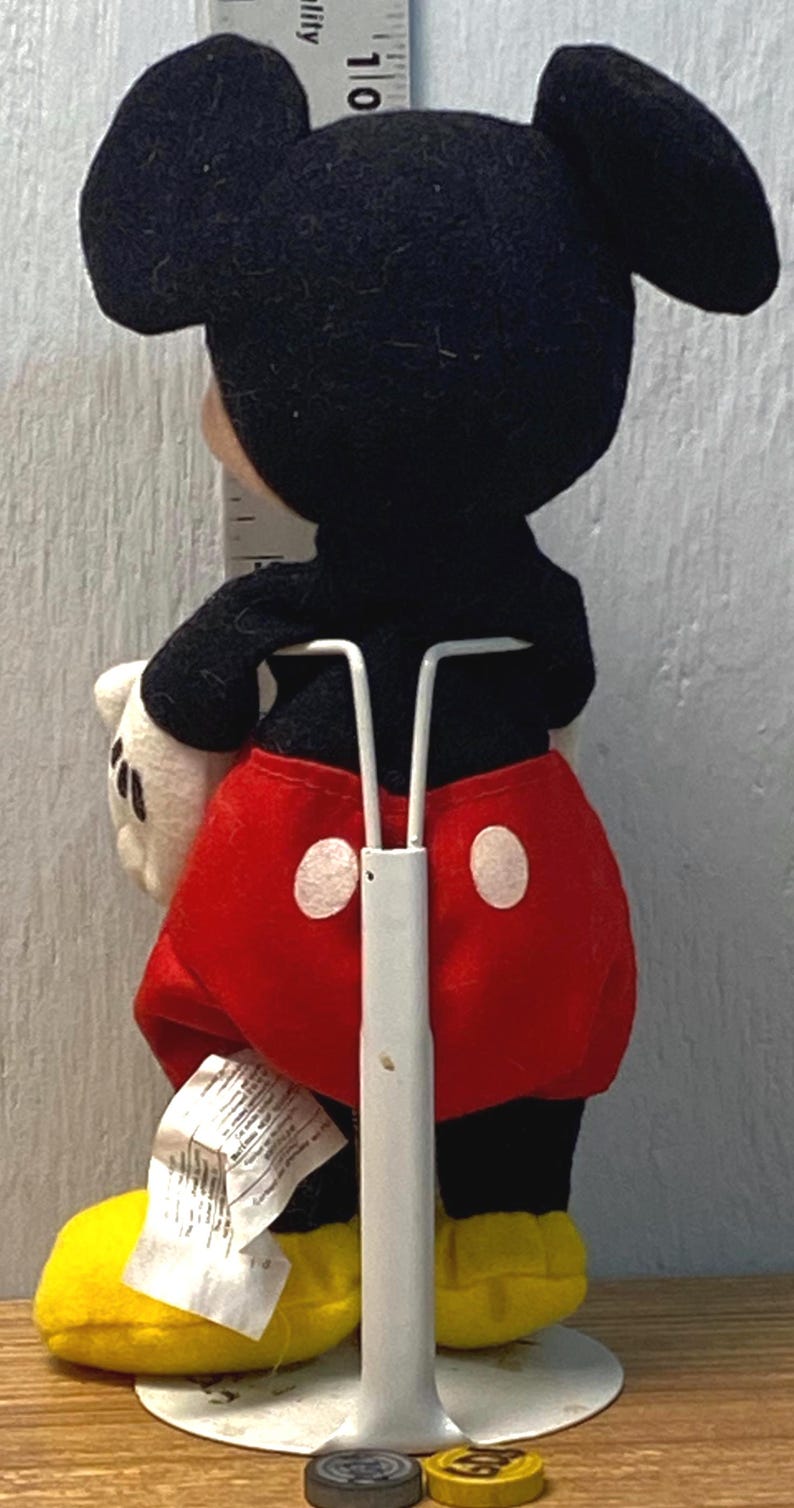 Puede incluir: Un peluche de Mickey Mouse, visto desde atr&aacute;s, con orejas negras, cuerpo negro, pantalones cortos rojos con botones blancos y zapatos amarillos. El juguete est&aacute; sobre un soporte blanco. Una regla es visible en el fondo.