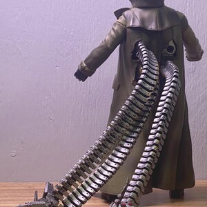 May include: A Dr. Octopus action figure, viewed from the back, wearing a long olive green coat. The figure has four articulated mechanical tentacles extending from the back. The tentacles are silver and gray with detailed mechanical components.