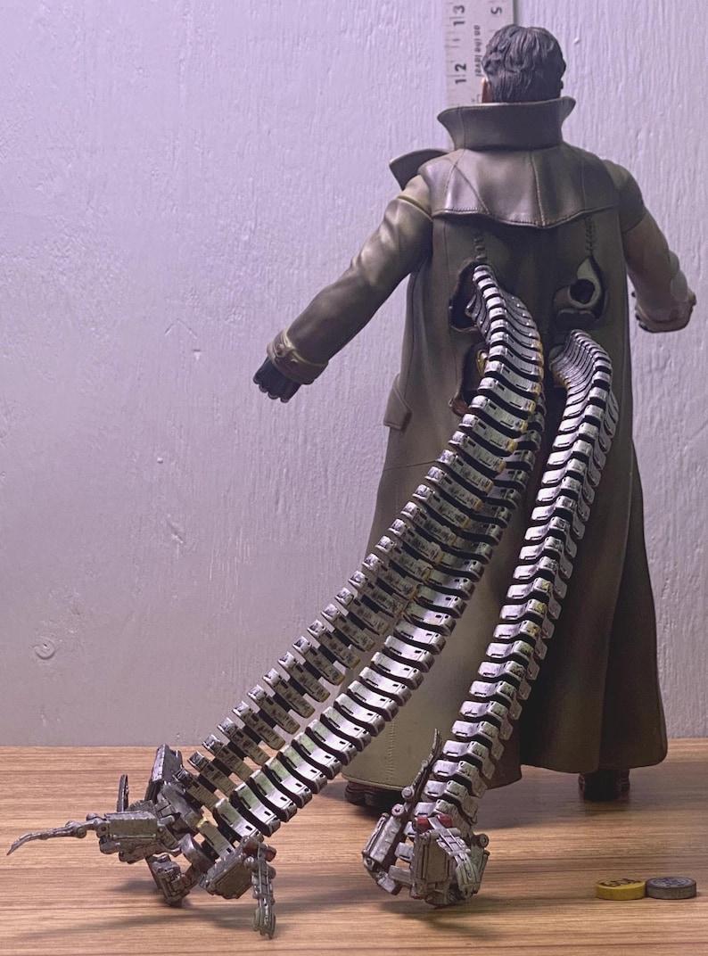 May include: A Dr. Octopus action figure, viewed from the back, wearing a long olive green coat. The figure has four articulated mechanical tentacles extending from the back. The tentacles are silver and gray with detailed mechanical components.