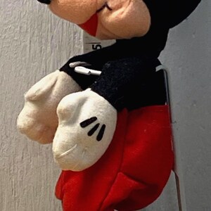 Puede incluir: Un peluche de Mickey Mouse, con cabeza y orejas negras, cara beige, guantes blancos, pantalones cortos rojos y zapatos amarillos. El juguete est&aacute; colocado junto a una regla, con peque&ntilde;os objetos redondos en la base.