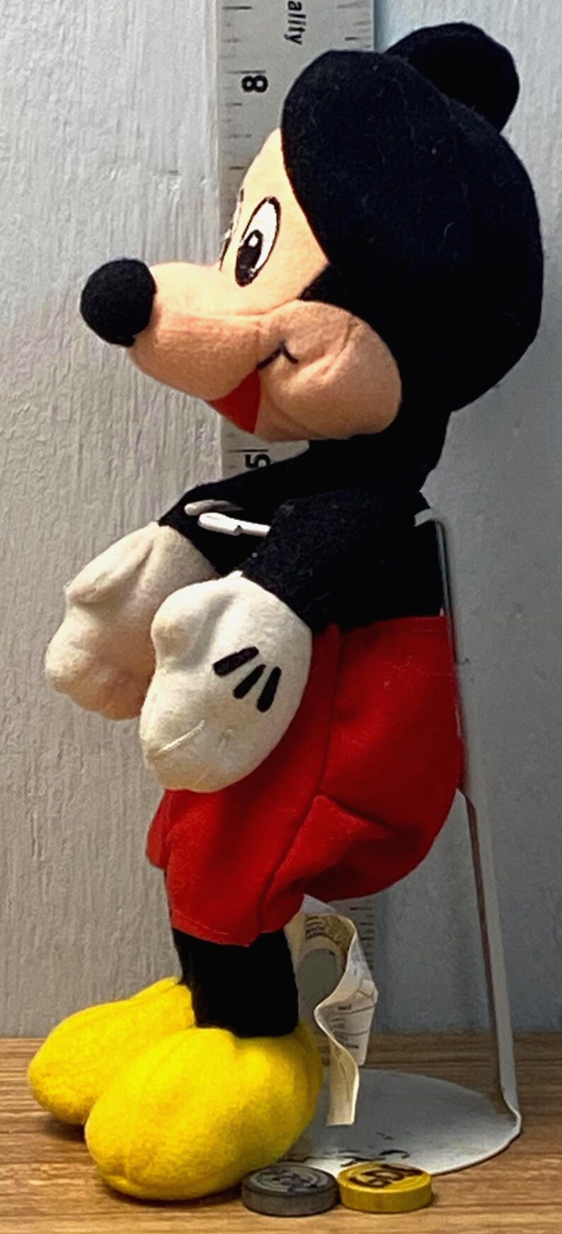 Puede incluir: Un peluche de Mickey Mouse, con cabeza y orejas negras, cara beige, guantes blancos, pantalones cortos rojos y zapatos amarillos. El juguete est&aacute; colocado junto a una regla, con peque&ntilde;os objetos redondos en la base.