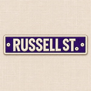 Pode incluir: Um emblema retangular bordado com um fundo roxo profundo e o texto branco RUSSELL ST. numa fonte negrito sem serifa. O emblema é contornado por uma borda espessa costurada de cor clara e inclui dois pequenos ilhós circulares.