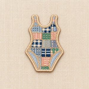 Puede incluir: Un parche bordado con forma de bañador de una pieza que presenta un diseño de patchwork con varios patrones geométricos en tonos azul, verde y melocotón, todo ello bordeado con un hilo dorado metálico grueso sobre un fondo de tela texturizada.