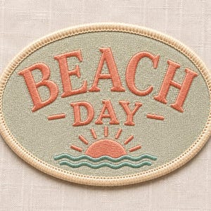 May include: An oval-shaped embroidered patch featuring a sage green background with a cream-coloured stitched border. The text BEACH DAY is embroidered in coral orange, positioned above a coral orange sun rising over teal blue wavy water lines.