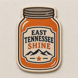 May include: An embroidered patch shaped like a mason jar with an orange body and a brown lid. The center features a cream-colored label with the dark blue text EAST TENNESSEE SHINE, a dark blue mountain silhouette, and a small orange star below.