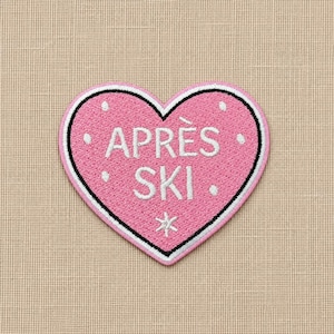 May include: A heart-shaped embroidered patch in pink with a black and white border. The text APRÈS SKI is stitched in white in the center, surrounded by small white dots and a white snowflake symbol at the bottom point of the heart shape.