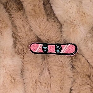 May include: A pink and black embroidered snowboard patch with blue bindings. The patch has a black outline and is set against a fluffy, light brown background.