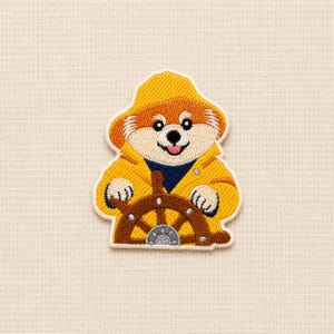 May include: An embroidered patch featuring a bear with light brown fur, wearing a yellow rain hat and a matching yellow raincoat over a dark blue shirt. The bear is holding a brown wooden ship steering wheel with a grey centre detail.