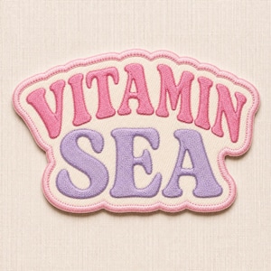 May include: An embroidered patch with a cream-coloured background and a scalloped pink border. The text VITAMIN is stitched in pink, and the word SEA is stitched in lavender. The letters have a rounded, bold font style throughout the design.